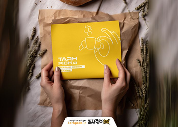 دانلود موکاپ بسته‌بندی نان باگت در دست با پس‌زمینه پارچه‌ای و گندم